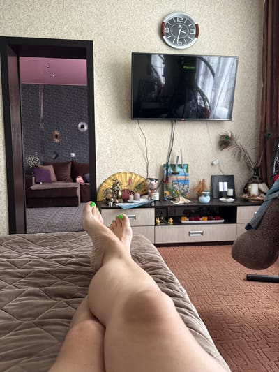 This is an indoor photograph with a person's legs in the foreground, resting on a quilted bed or sofa. The legs are crossed at the knee, and the toenails are painted a bright green color. The person appears to be relaxed.
In the background, a television is mounted on a wall with textured wallpaper. To the right of the television is a tall cabinet with shelves displaying various decorative items, including a peacock feather, candles, seashells, a small painting of a beach scene, and an ornate fan. A clock with a dark face and white numerals is visible on the wall to the left of the television.
Below the television and cabinet is a framed mirror that reflects the room. The reflection shows more of the room's furnishings, including a couch with brown and purple cushions. The overall impression is of a living room or bedroom. The lighting suggests it is daytime, but the specific time is not clear from the clock's position. There are no external weather cues visible. The location is identified as Kamen-na-Obi, Russia.