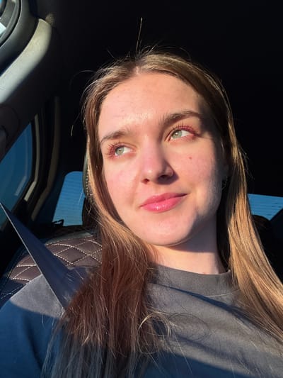 A young woman with light brown hair and green eyes is sitting in the passenger seat of a car, looking to her right. She is wearing a dark gray t-shirt and has pink lipstick on. The sun is shining through the window, casting a shadow across her face and the interior of the car. The car's interior is dark, with a dark gray seat visible behind her and a seatbelt across her chest. The location is implied to be Ustinovo, Russia, although no specific landmarks are visible.