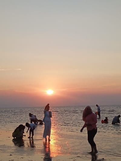 The image captures a sunset scene at a beach or tidal flat in Tritunggal, Indonesia. The sky transitions from light blue-grey at the zenith to vibrant orange and pink hues near the horizon, where the sun is visible as a bright orange orb, positioned slightly left of center. The sun's reflection creates an illuminated path across the shallow water and wet sand towards the foreground.
Approximately ten to twelve individuals are present. In the foreground-left, one person in yellow attire is crouched, alongside a small child in a white t-shirt, both observing the sand or water. Slightly right of them, a woman in a light-colored long dress and a head covering stands upright, holding a dark object, possibly a phone, upwards as if photographing or recording. To her right, a woman in a pink top, dark pants, and a head covering stands, facing slightly left, with one foot partially elevated.
Further into the mid-ground and background, several figures are dispersed across the shallow water. Some are standing, while others are bent over or crouched, seemingly engaged in activities within the water. One individual in the background-right is observed near a red or orange inflatable ring. The water is calm with minor ripples, reflecting the ambient light and the sun's glow. The wet sand in the immediate foreground also reflects the sky and light.