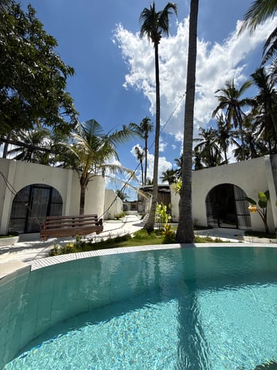 The image captures a tropical scene in Gubug, Indonesia, featuring a swimming pool adjacent to a white, modern building with distinctive arched windows. Palm trees are interspersed throughout the frame, contrasting with the blue sky and scattered clouds above. The water in the pool shimmers with reflected sunlight. A wooden bench or decorative feature is attached to the building's exterior, adding a rustic touch to the modern architectural design. The scene appears peaceful and relaxed, conveying a sense of tropical leisure.