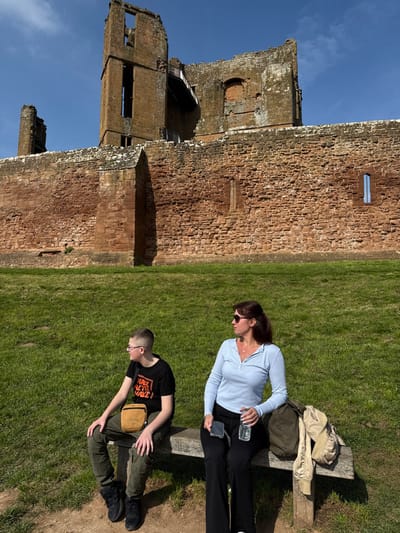 A daytime image captured in Kenilworth, Warwickshire, United Kingdom, features two individuals seated on a wooden bench in the foreground, with Kenilworth Castle ruins prominent in the background under a clear blue sky.
The scene shows a grassy embankment leading up to a large, reddish-brown stone wall, part of Kenilworth Castle. Above this wall, additional ruined castle structures are visible, including a tall, partially collapsed tower with exposed interior levels and window openings. The stone shows weathering and some light-colored accumulation along its upper edge.
On the wooden bench, positioned in the lower-middle of the frame, sits an adult female on the right and a younger male on the left. Both are looking to their left, away from the camera's direct line of sight.
The female wears sunglasses, a light blue long-sleeved collared shirt, black trousers, and holds a clear plastic water bottle in her right hand. A dark-colored smartphone is visible near her left hand. To her right, on the bench, rests a dark olive-green fabric bag and a light tan jacket.
The male wears glasses, a black short-sleeved t-shirt with orange and white text, dark olive-green cargo-style trousers, and black lace-up boots. He has a tan cross-body bag positioned across his chest. His hands are resting on his lap.
The ground directly in front of the bench is a dirt patch, transitioning into a vibrant green grassy area that extends towards the castle wall. The lighting suggests a bright, sunny day, with distinct shadows cast by the subjects and objects.