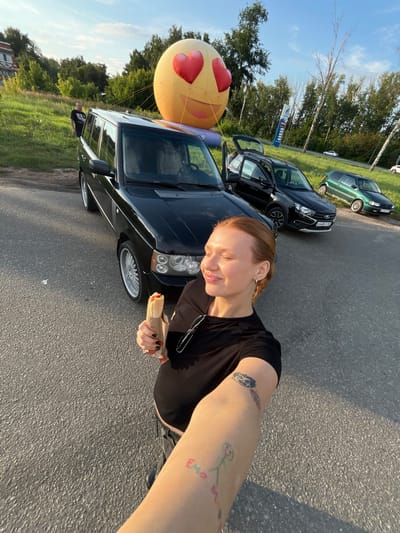 A woman with red hair, wearing a black t-shirt, is taking a selfie outdoors. She is holding a food item wrapped in paper in her right hand. She has several tattoos on her left arm. In the background, a black Range Rover is parked, and behind it, a large yellow inflatable emoji with heart-shaped eyes is visible. Other cars are parked further in the background, along with trees and a building. The scene appears to be a street or parking area, likely in Tri-Izby, Russia, given the context of the prompt. The sky is blue with some clouds, and the lighting suggests it is daytime, possibly late afternoon.