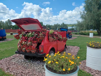This is a medium shot, eye-level, outdoor photograph captured on a cloudy day in the city of Kovalivka, Ukraine. The foreground features a red, vintage car with its trunk open, filled with numerous potted flowers of various colors, including red, pink, white, and green. The car is positioned on a bed of gravel. To the right of the car, a large white planter holds a cluster of yellow flowers. A second, larger planter filled with yellow flowers sits further to the right. The background consists of a lush green lawn, trees, a body of water, and a partly cloudy blue sky. A blue vehicle, also containing potted flowers, is partially visible to the left of the red car. Several small, black lampposts are scattered across the lawn.