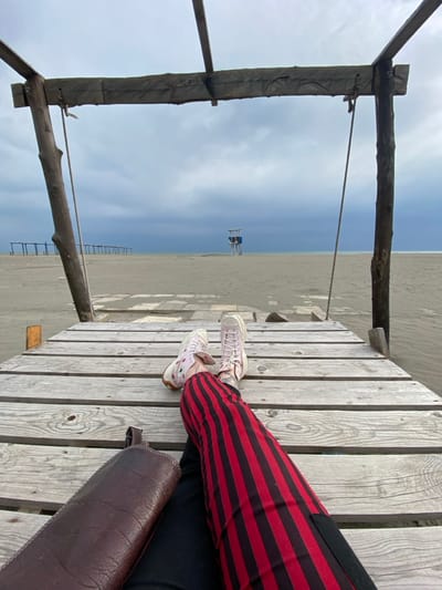 The image depicts a first-person view from a weathered wooden platform on a sandy beach in Ulcinj, Montenegro, under an overcast sky. In the immediate foreground, a person's legs are extended, clad in red pants with vertical black stripes and light pink high-top sneakers. A dark brown, textured bag rests on the left thigh. A rudimentary wooden frame structure, consisting of vertical posts and a horizontal crossbeam, partially frames the upper left of the scene, with a rope hanging from the left post. Beyond the platform, a wide expanse of light brown sand stretches towards the horizon. A blue and white lifeguard tower stands centrally on the distant beach, slightly to the right. Further to the left on the sand, a linear series of dark wooden posts connected by crossbeams is visible. The background consists of a calm, dark blue-grey sea meeting a light grey, heavily clouded sky.