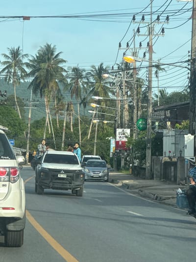 The image depicts a street scene in Baan Tha Po, Thailand, characterized by a clear, bright sky and lush palm trees lining the roadside. The road is paved and shows signs of traffic, with several vehicles visible, including a white Toyota Fortuner on the left, a silver Mitsubishi Triton pickup truck in the center, and a silver sedan further down the road. The Mitsubishi Triton has a license plate that reads "WU 1406".

There are people in the scene, with two individuals standing on the back of what appears to be a motorcycle or a similar vehicle behind the Mitsubishi Triton. One person is wearing a plaid shirt, and the other a blue shirt. Another person is partially visible in the driver's seat of the pickup truck.

The environment is a mix of natural and urban elements. Numerous utility poles with a dense network of electrical wires dominate the background. Roadside structures include buildings and a stone wall. Several signs are visible, including a green circular "Chang" beer logo, and other signs with Thai script. A street light with an orange glow is also present. The overall impression is of a busy tropical street with typical infrastructure.