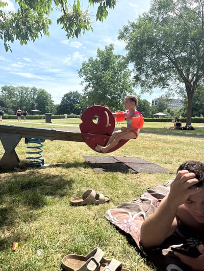 The image depicts a bright, sunny day at an outdoor park or playground in Giessen, Germany. In the foreground, a young girl, wearing a colorful swimsuit and orange arm floaties, is engaged in playing on a red seesaw. She appears to be mid-action, leaning forward on the seesaw. Beside her, to the right, a person is lying on a patterned blanket, with their hand touching their head, possibly relaxing or napping in the sun. Two pairs of light brown sandals are visible on the grassy ground near the blanket.

In the background, other park-goers can be seen enjoying the outdoors. Some are walking on a paved path, while others are seated or lying on the grass. Trees provide shade, and a modern building with multiple windows is visible in the distance, suggesting an urban park setting. The sky is a clear blue with scattered white clouds, indicating fair weather. The overall atmosphere seems relaxed and leisurely, with people enjoying a warm day at a public recreational space. No visible text is present in the image.