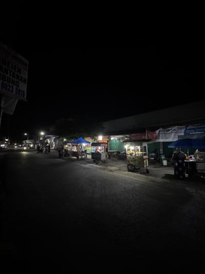 This image captures a street scene at night in Alian, Indonesia, likely a commercial area with several food stalls lining the right side of the road. The street is paved and appears wet, reflecting the artificial lights from the stalls and passing vehicles. On the left side of the road, a sign is partially visible with text that appears to be in Indonesian, including numbers that might be a phone number.
Several food carts are set up along the sidewalk on the right. One cart, prominently displayed, has a sign that reads "TUYO OYE" and appears to be selling fried food. Another cart under a blue umbrella has a person in a dark patterned dress standing next to it, seemingly attending to customers or preparing food. Other carts are visible further down the street, some with awnings providing shelter.
A few motorcycles are parked near the stalls, and a person is seen on one of them. The overall atmosphere is that of a bustling night market, with the lights from the stalls illuminating the immediate surroundings against the dark night sky. The buildings behind the stalls are dimly lit, and trees can be seen in the background, adding to the urban yet slightly rural feel of the location. The presence of multiple food vendors suggests a local gathering spot for evening snacks and meals.