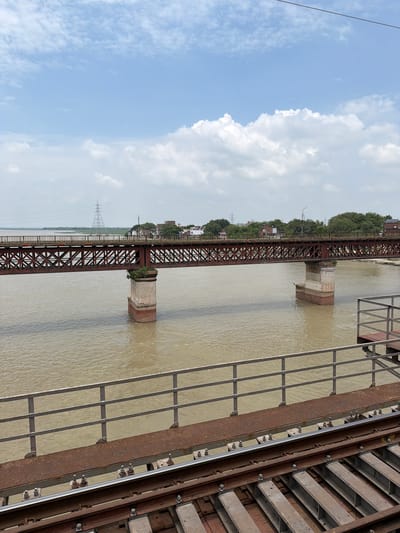 A horizontal, rusted metal bridge spans across a wide, muddy river. The bridge has a lattice-like structure and is supported by two cylindrical brick pillars, one of which has vegetation growing on top. On the far bank of the river, there are trees, buildings, and a tall electricity pylon. In the foreground, railroad tracks run along a rusty metal walkway with a railing. The sky above is cloudy and blue. The scene appears to be in Shuklaganj, India, given the characteristic architecture and environment. There are no people visible in the image.