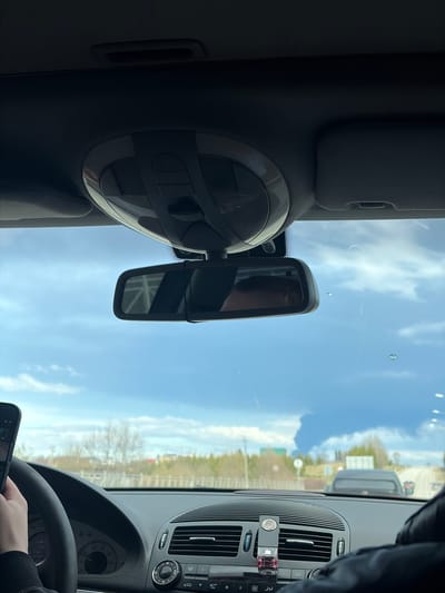 The image depicts an interior view from the driver's seat of a car, presumably a Mercedes-Benz based on the dashboard design. The dashboard features a central air vent system with attached air fresheners. The rearview mirror is prominent, and the car's interior lights fixture is visible. Outside the windshield, the sky is blue with scattered clouds. The environment appears to be a road with trees and vegetation on the sides, possibly in a suburban or rural area. The phone in the driver's hand may be recording a video, but this is not confirmed. The location, Yugo-Kamskiy, Russia, is not identifiable within the image.