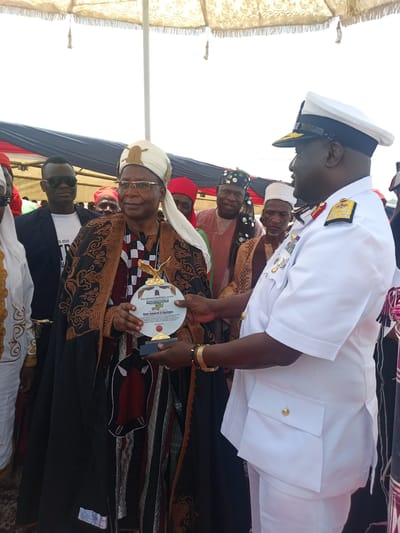 The image depicts a formal event in Dilpurtu, Nigeria. An elderly man, adorned in traditional African attire, including a white turban, embroidered robe, and glasses, is holding an award. The award appears to be circular with an eagle emblem. A man in a white naval uniform, complete with shoulder boards, gold braid, and medals, presents the award. Several individuals in traditional Nigerian garments surround them. An awning or shade structure is visible in the background.