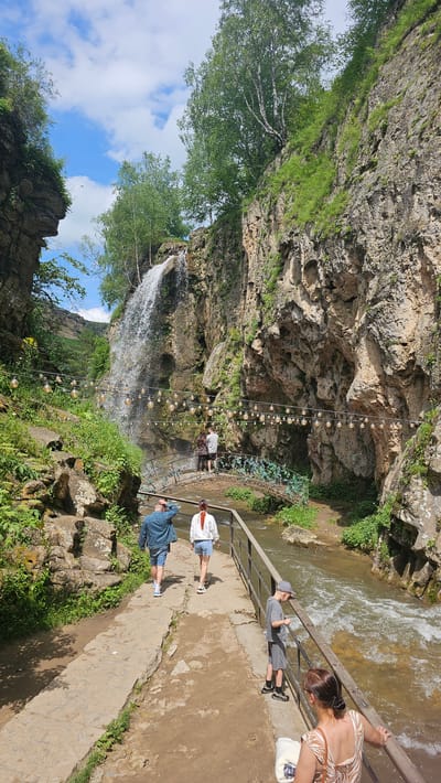 The image depicts a scenic natural landscape with a waterfall cascading down a rocky cliff face. A river flows through the scene, with a pedestrian bridge spanning across it. String lights are strung across the gorge, adding a decorative element. Several people are present, walking along a path and on the bridge. The environment is characterized by lush greenery and rugged rock formations, under a bright blue sky with scattered clouds. The location is identified as Красный Курган, Russia.