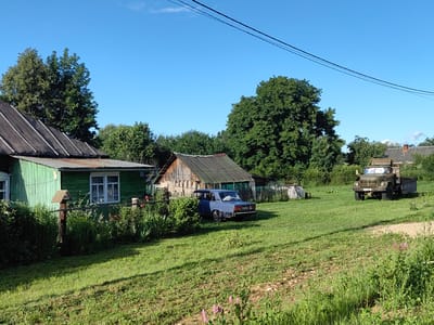 The image shows a rural scene in the city of Квант-88 Снт, Russia. In the foreground, a lush green lawn slopes gently downwards, with patches of dry earth and scattered wildflowers. The middle ground features a green wooden house with a metal roof, a fence, and a bird feeder on a wooden post. Next to the house is a wooden shed with a corrugated metal roof. A blue sedan is parked in the yard. In the background, a large green military truck is parked on a dirt road. Tall trees and dense foliage surround the property, and a clear blue sky with a few wispy clouds forms the backdrop. There are no people visible in the image.