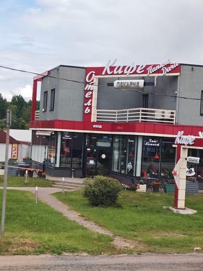 The image shows a two-story building with a modern architectural design, featuring a grey facade and red trim. The building houses a cafe and a bakery, indicated by large, illuminated signs. The main sign above the cafe reads "Кафе "Плантация Дара" (Cafe "Plantation of Dara") in stylized white lettering, with "ПЕКАРНЯ" (Bakery) written below. Another vertical sign, also labeled "ПЕКАРНЯ", stands to the right of the cafe entrance. The cafe has large glass windows displaying tables and chairs inside, where a few people are seated. The bakery section is also visible through the windows, with displays of baked goods. The exterior of the cafe is adorned with potted flowers and outdoor seating areas. The building is situated in a semi-urban or suburban environment, with a paved pathway leading to the entrance, surrounded by green grass and trees in the background. The sky is overcast. The building is located in the city of Dolgii Most, Russia.