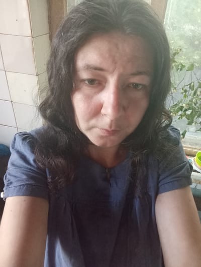 A woman with dark, wavy hair is looking at the camera with a neutral expression. She is wearing a blue short-sleeved shirt. The background shows a tiled wall on the left and a window with green foliage on the right. The scene appears to be indoors, likely a domestic setting. The image is an over-the-shoulder selfie. No specific location in Berezivka, Ukraine, can be identified from the visual information.
