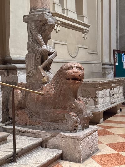 The image displays a sculpted stone ensemble located within the Museo della Cattedrale di Ferrara, in Ferrara, Italy. Central to the frame is a reddish-brown stone lion figure, depicted in a prone position, facing right with its mouth agape, resting on a textured stone base. Above the lion, a light reddish-brown stone figure of a male (an Atlante) is carved in a crouched stance, supporting the circular base of a pinkish-red column with both hands. His right leg is bent, and his left leg is extended.
To the immediate left foreground, several light-gray stone steps ascend. A dark metal stanchion featuring a brass-colored horizontal rod extends diagonally from the lower left, acting as a barrier in front of the sculpture. The floor is composed of polished, reddish-brown and off-white checkered tiles.
In the mid-ground to the right of the lion, a large, rectangular, light-colored stone sarcophagus or tomb is visible, adorned with intricate bas-relief carvings. The background features a light-beige architectural wall detailed with classical moldings, pilasters, and recessed panels. A dark green information board with several white papers affixed to it is partially visible in the far right background. The scene is lit by ambient indoor illumination.
