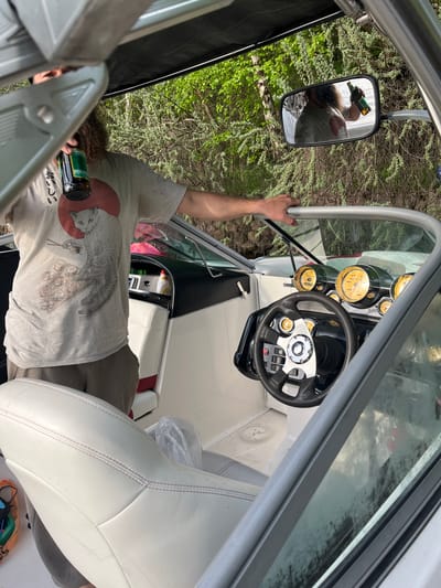The image shows the interior of a boat, possibly a motorboat, with a fair-skinned individual holding a dark-colored bottle. The person is wearing a light-colored t-shirt with a graphic design. A mirror on the side of the boat reflects the person's face and the bottle, along with the surrounding natural environment. The boat's dashboard features various gauges and a steering wheel. The boat is white with interior upholstery. The environment outside the boat appears to be a forest or wooded area. It is not possible to confirm the location as Пятница, Russia from the image alone.