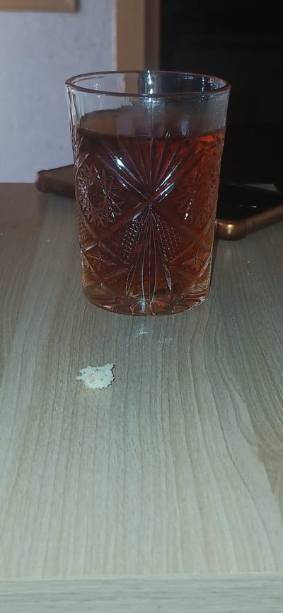 A glass of amber liquid sits on a wooden table. A small, pale, irregular object with pink specks is on the table in front of the glass. A smartphone is partially visible behind the glass. The scene appears to be indoors, possibly in a home in Khyrdalan, Azerbaijan.