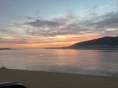 The image displays a panoramic, horizontal view of the Minho River estuary during either sunrise or sunset, observed from a vehicle within Caminha, Portugal. The sky presents a gradient from light blue and grey overhead to prominent bands of orange, pink, and yellow light across the horizon, containing thin, horizontally elongated clouds. The vast body of water is calm, reflecting the sky's colors with subtle surface ripples. On the far left horizon, a low-lying landmass exhibits faint lights. To the right, the dark, silhouetted mass of Monte Santa Tecla (A Guarda, Spain) dominates, its base meeting the water. Numerous small, bright lights from a settlement are visible along its lower slopes. The immediate foreground, comprising the bottom fifth of the frame, is a blurred, dark grey, flat surface consistent with a road barrier or bridge railing. A dark, curved object, likely part of a vehicle's interior, is visible in the lower-left corner. No people or specific interactions are depicted.