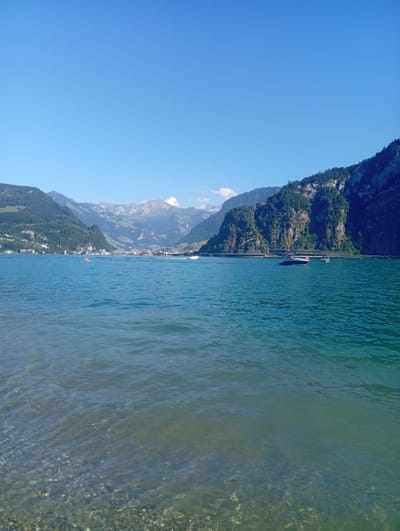 The image captures a tranquil scene on Lake Lucerne, looking towards the town of Hergiswil in the canton of Nidwalden, Switzerland. The foreground is dominated by the clear, shallow waters of the lake, revealing a pebble-strewn bottom. Gentle waves create ripples on the surface.

In the mid-ground, the expansive blue water extends towards the opposite shore. Several boats are visible: a small red inflatable craft with at least two people aboard is to the left of center, while a sleek motorboat with an orange life vest visible on its stern is positioned to the right of center. Another white boat is partially visible further to the right. A distinct, low bridge or causeway stretches across the water in the distance, connecting the shores.

The background is a dramatic panorama of mountains. The left side features a green, tree-covered slope leading up to rugged, layered mountains. In the center, a cluster of buildings representing the town of Hergiswil is nestled at the base of the mountains, with the higher peaks beyond showing patches of snow and rock. The right side of the background is dominated by a steep, dark, and craggy mountain face densely covered in trees. The sky above is a vast, clear, and brilliant blue. The overall impression is one of a serene summer day in a picturesque alpine setting.