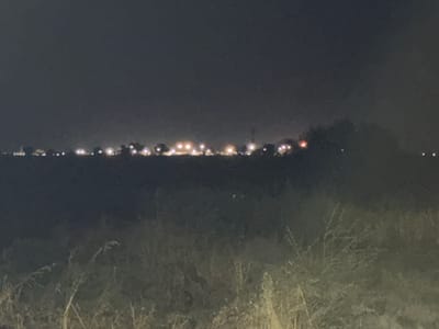 The image depicts a landscape at night, with a faint line of lights in the distance, suggesting a settlement or town. The foreground is dominated by dry, tall grass and vegetation, rendered in dark, muted tones due to the low light conditions. The sky is dark and overcast, with no visible stars. The distant lights appear as scattered points of illumination, some brighter than others, and are silhouetted against the dark horizon. There are no discernible people or specific structures visible, only the general impression of a rural or semi-rural area at night with lights from a distant source. The overall atmosphere is dim and somewhat blurry, characteristic of a nighttime photograph taken with limited light.