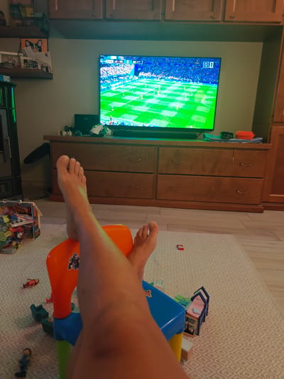 The image depicts a domestic living room setting, likely in an apartment or house, where a person is relaxing and watching television. The television screen displays a live soccer match. In the foreground, a pair of bare feet are propped up on a multi-colored plastic toy chair. Scattered around the toy chair are various small toys, including miniature figures and building blocks, suggesting the presence of children or a child-friendly environment. The furniture includes a wooden dresser with multiple drawers, and above it, a shelf with decorative items and framed pictures. The room is illuminated by ambient lighting and the glow from the television. The floor is covered with a light-colored rug. There is no information or visual evidence present in the image to suggest or confirm the location in the city of Carmagnola, Italy.