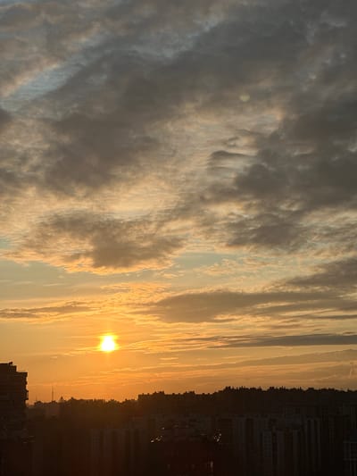 This is a vertical, high-angle photograph of a city skyline at sunrise. The sun, a bright, white-yellow orb, is positioned in the upper left quadrant, partially obscured by thin, elongated clouds. The sky transitions from a pale yellow near the horizon to a dusty gray and light blue higher up. Wispy clouds are scattered throughout, with some appearing denser and darker.
The lower half of the image is dominated by the silhouetted cityscape. Buildings of varying heights, including apartment complexes and what appear to be industrial structures, form a dark, undulating line across the horizon. A tall, thin tower with a geometric structure at its apex is visible in the mid-ground. The foreground features the dark shapes of more buildings, including a larger, multi-story structure on the far left. There are no visible people or any discernable actions or interactions occurring within the scene. The environment suggests an urban setting at the beginning of the day, with a focus on the interplay of light and shadow on the clouds and architecture. The location is Feklistovo, Russia.
