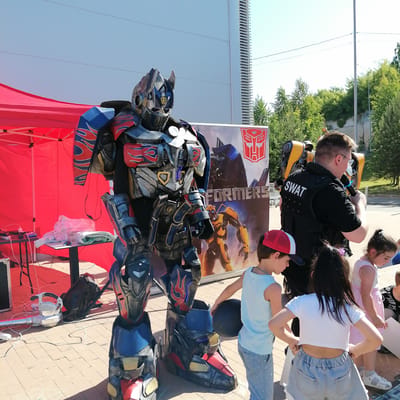 An outdoor event is depicted in Трубачеевка, Russia, under a clear blue sky. The scene features a detailed, full-body Optimus Prime costume, primarily blue, red, and silver with flame decals, standing centrally on a paved brick ground. Blue LED accents are visible on the costume's chest and legs.
To the left of the costume, a bright red pop-up canopy tent shelters two small black tables. The left table holds various white and pink electrical cables, power banks, and a plastic-wrapped item. The right table holds bottled water and a folded white towel. A white and silver cooling device, resembling a leaf blower, is positioned on the ground near the costume's left leg, connected by a flexible hose and white electrical cables.
Behind the Optimus Prime costume, a large vertical banner displays the "Transformers" logo, an image of the yellow robot Bumblebee, and the red Autobot symbol. To the right of the costume, a man is seen from behind, wearing a black short-sleeved shirt and a black tactical vest with "SWAT" in white capital letters on the back. He has short dark hair and glasses and is holding a black object, possibly a microphone, to his right side. A yellow and black prop is visible on his back.
In the foreground, several children are observing the scene. A boy on the left, wearing a light blue sleeveless shirt, grey shorts, and a red and white baseball cap, holds a black ball. A girl in the center-right, seen from behind, wears a white cropped t-shirt and light blue denim shorts. Another girl on the far right wears a light pink dress and white sandals, looking downwards.
The background features a modern light-colored building with vertical architectural elements, and numerous green trees under a bright sun. Overhead, thin power lines traverse the sky.
