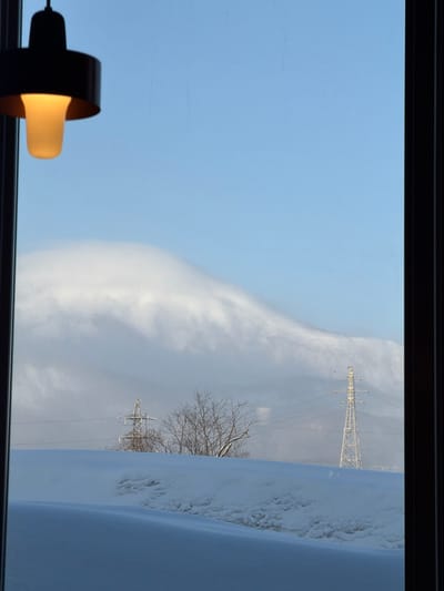 A window frames a winter scene. Bare tree branches and a transmission tower are partially obscured by snow drifts. Wispy clouds and a light blue sky fill the background. In the lower left corner, a lamp with a black shade and glowing bulb is visible. The window frame is black.
