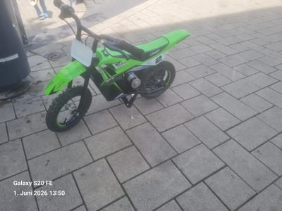 The image shows a Razor SX McGrath electric dirt bike parked on a paved pedestrian area. The bike is green and black, with a white number plate on the front. The seat is also green, and the rear fender has the Razor logo and the number "2". The bike is positioned on a gray paved surface composed of rectangular blocks. In the upper-left corner, there is a portion of a black cylindrical object, possibly a bollard or part of a lamppost. Partial figures of people are visible in the far upper-left. The location is likely within an urban setting within the city of Bingen am Rhein, Germany. The date and time stamp at the bottom of the image indicates the picture was taken on June 1, 2026, at 13:50.