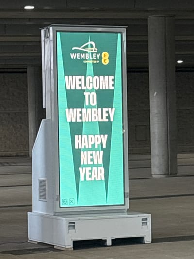 A brightly lit, vertically oriented digital display screen is the central subject, featuring a vibrant green background with prominent white text and graphics. The screen is mounted within a light gray frame, which appears to be part of a larger fixture, visible with a ventilation grille on its lower right side.
The display shows a welcoming message. In the upper left corner, the Wembley logo, an abstract 'W' design, is visible, accompanied by the words "CONNECTED BY EE" and the orange EE logo. The main message, stacked vertically and readable from top to bottom, states "WELCOME TO WEMBLEY HAPPY NEW YEAR." The text is large, bold, and white, set against the green background, with subtle lighter green triangular shapes extending from the text. In the bottom right corner of the screen, small icons, including a camera symbol and a square, are visible.
The screen is set against a dark, indistinct background, likely a wall or structural element, suggesting an indoor or covered public area. The lighting in the background is dim compared to the illuminated screen. The overall scene indicates a festive and welcoming atmosphere, consistent with a public venue in Wembley, United Kingdom, around the New Year period.