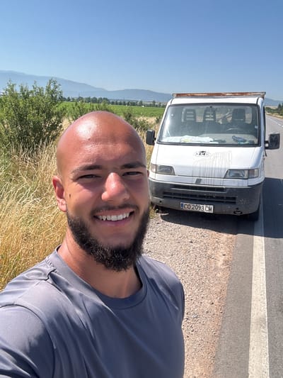 This media file is a portrait-oriented selfie taken outdoors on a sunny day. The primary subject is a bald, dark-skinned man with a beard, smiling and looking at the camera. He is wearing a grey t-shirt. In the background, to the left of the man, is the front of a white flatbed truck. The truck has a dark grey bumper and a license plate that reads "CO 2093 CM". The background also shows a rural landscape with green fields and distant hills under a clear blue sky. The sun appears to be high in the sky, indicating it is likely midday. The ground near the man is covered in dry grass and some bushes. The road is visible in the upper right corner of the image. The overall impression is one of a casual, outdoor moment, possibly during travel or a break in activity.