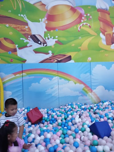 In Talas City, Kyrgyzstan, a boy and a girl stand in a ball pit. The boy, wearing a striped t-shirt, looks to his left while the girl, wearing a pink shirt, looks at the balls in front of her. The ball pit is filled with a variety of colored balls, including pink, blue, white, and green. A red cube and a blue triangular prism are also visible within the ball pit. Behind the ball pit, a backdrop depicts a whimsical candy-themed landscape with rolling green hills, chocolate bars, and candy canes, as well as a blue sky with a rainbow and clouds.