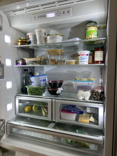 The interior of a stainless steel refrigerator is open and illuminated by several white LED lights. The refrigerator is in a kitchen in Rye, New York, located at the coordinates provided. The top shelf is lined with containers of food, including yogurt, cottage cheese, sour cream, and a jar of pickled green beans. The second shelf contains jars of food, including pickled peppers, a container of sour cream, and a jar of salsa. The third shelf contains food containers, including a container of caviar, various containers of unidentified food, and a clear container with brown food items. The fourth shelf contains a clear container with food items, several containers of unidentified food, and a container with a blue lid and the word "dairy" printed on it. The fifth shelf contains two large containers of unidentified food, a container of blue berries, and a container of asparagas. The sixth shelf contains a clear container of unidentified food, a red container of unidentified food, and a container of asparagas. The seventh shelf contains a container of unidentified food, a container of dairy cheese, and two containers of unidentified food. The bottom shelf contains a container of green peas. The refrigerator has a white plastic interior. There is no visible person or any action taking place in the image.