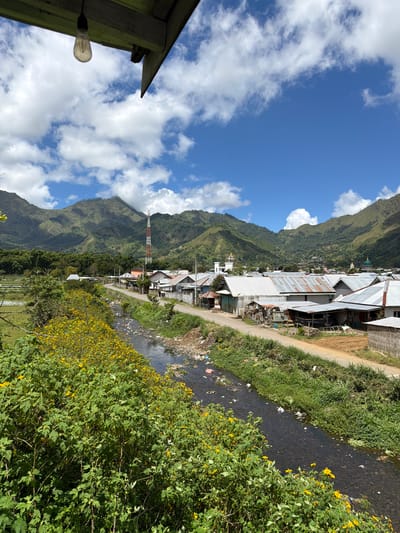 The image depicts a rural landscape in Sembalun Bumbung, Indonesia. A stream flows through the scene, bordered by vegetation including bushes with yellow flowers. A dirt road runs parallel to the stream, alongside buildings constructed with metal roofs. In the distance, a mountain range rises with patchy cloud cover. A red and white communication tower stands among the buildings. The sky is blue with scattered clouds. Debris litters the stream.