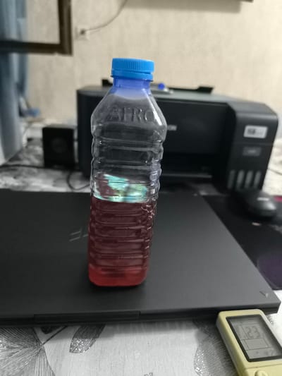 An indoor, eye-level, medium shot captures a partially filled plastic bottle with a blue cap. The bottle contains a red liquid with a layer of lighter colored liquid on top. It is placed on a dark laptop, which is positioned on a table covered with a patterned tablecloth. In the background, a black printer is visible, along with a small black speaker. The scene is well-lit, with shadows cast by the objects. There are no people present in the image. The location is not discernible within the image, and therefore its presence in Kokand, Uzbekistan, cannot be confirmed.