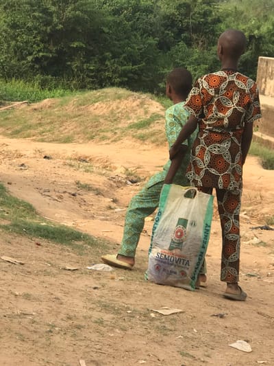 Two children carry a bag of "Golden Penny Semovita" brand semolina. The child in front wears a green patterned outfit, and the child behind wears an orange and brown patterned outfit. They both wear slip-on shoes. They walk along a dirt path, with a grassy slope on the left and a dirt surface on the right. A building is visible in the background.