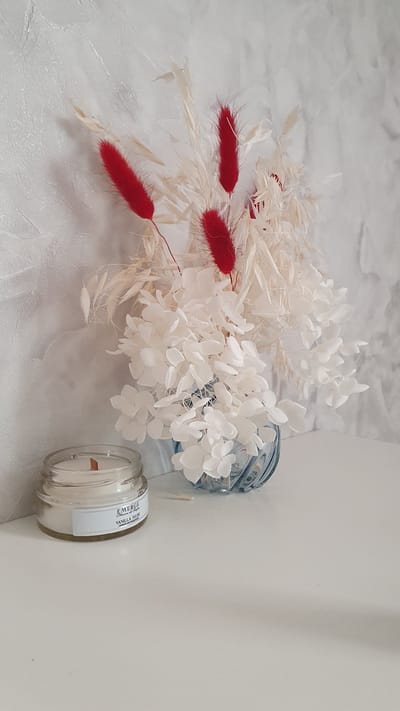 A small, glass jar candle with a wooden wick sits on a white surface in front of a textured grey wall. The candle has a white label with blue text that reads "EMERGE Vanilla Musk Candles and Potpourri." To the right of the candle is a blue, ribbed glass vase containing a dried floral arrangement. The arrangement features white, delicate flowers and tall, creamy-white feathery stalks, interspersed with several bright red, fuzzy elements. The overall scene appears to be an indoor still life, likely a decorative display. There is no indication of the city of Aprelevka, Russia.