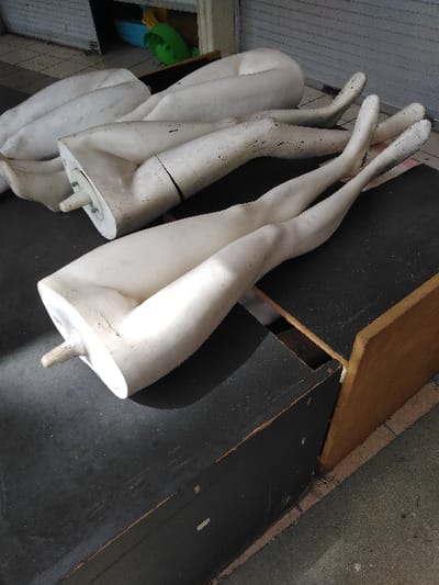 The image displays an outdoor or semi-outdoor storage area in Elverum, Norway. Several disarticulated white mannequin leg sections are present, arranged horizontally and piled on dark, flat surfaces, consistent with the tops of wooden crates or display units. The mannequin components exhibit signs of wear, including surface dirt and discoloration. In the background, a light-colored roller shutter door is partially raised, revealing a glimpse of an interior space containing indistinct green and blue objects. The floor visible in the background is light-colored, possibly concrete or tile. No individuals are present, and no actions or interactions are depicted within the scene.