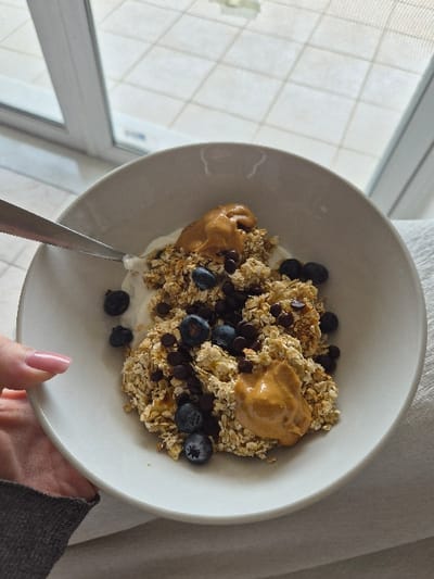 A human hand, featuring light pink manicured fingernails and clad in a grey long-sleeved garment, holds a light-colored ceramic bowl from the lower left. The bowl contains a base of white creamy substance, covered by light brown granola or oatmeal. Mixed within the granola are numerous dark blue blueberries and small dark brown chocolate chips. Three distinct dollops of light brown, viscous peanut butter are situated on top of the granola. A silver spoon is partially visible, resting within the bowl on the upper left side.

To the right of the bowl, a light-colored, textured fabric surface is present. In the upper background, a glass door or window frame is visible, separating the interior setting from an exterior space. Through the glass, an outdoor tiled surface composed of light-colored, square tiles arranged in a grid pattern is observed. The scene is situated within the Municipal Unit of Corinth, Greece.