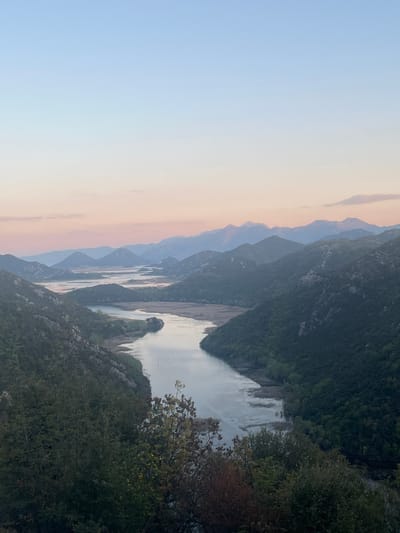 The image shows a mountain landscape in Rvaši, Montenegro. A river or lake winds through the scene, flanked by steep, forested mountains. The sky is a gradient of blues and pinks, indicating either sunrise or sunset. There is vegetation in the foreground, including trees and bushes. In the distance, layers of mountains are visible, receding into the horizon.