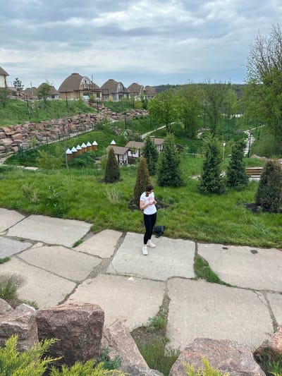 A woman stands on a concrete path in a landscaped area, possibly Zakharivka, Ukraine. She wears a white t-shirt, black pants, and white sneakers. A black bag sits near her feet. The path is constructed of irregularly shaped concrete slabs. To the left, a stone wall borders a grassy area with manicured evergreen trees and small, peaked-roof structures. In the background are buildings with rounded roofs. The sky is overcast.