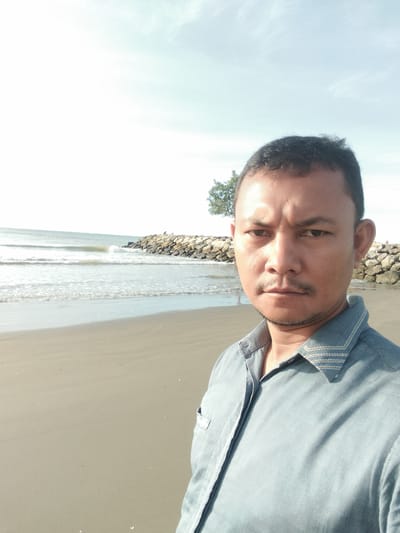 A man is taking a selfie on the beach in Blanglancang, Indonesia. The man is facing the camera, and he is wearing a blue collared shirt. The background of the image consists of a beach, the ocean, and a stone jetty. The sky is clear and blue.
