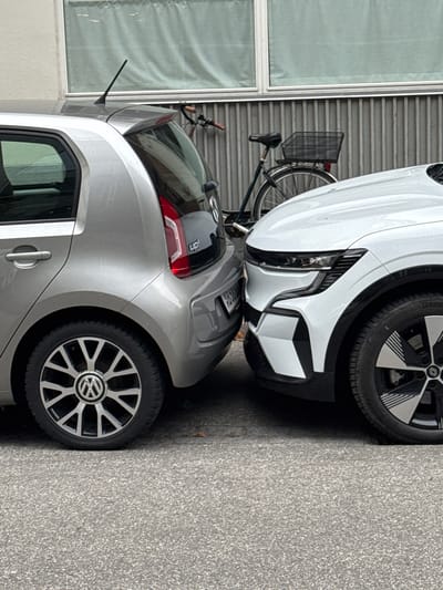Two cars, a silver Volkswagen Up! and a white Renault Megane E-Tech, are parked very close together on a street in Frederiksberg, Denmark. The silver car is on the left, and its rear bumper is visible, along with its "up!" badging. The white car is on the right, and its front bumper, headlights, and wheel are visible. The cars are parked on asphalt with a grey, ribbed building facade in the background. A bicycle with a basket is leaning against the building behind the cars.