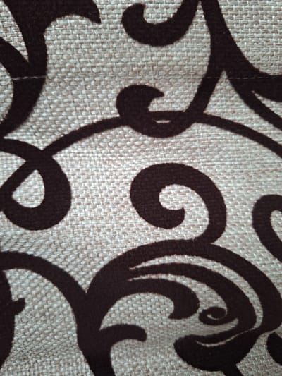 This is a close-up, high-angle shot of a textured fabric with a dark brown ornate scroll pattern. The fabric appears to be a neutral beige or light brown, with a visible woven texture. The pattern consists of thick, flowing lines that create swirling and looping shapes. The lighting suggests an indoor setting, possibly with overhead light casting subtle shadows. The image provides no information about the city of Новобурейский, Russia, or any specific location within it.