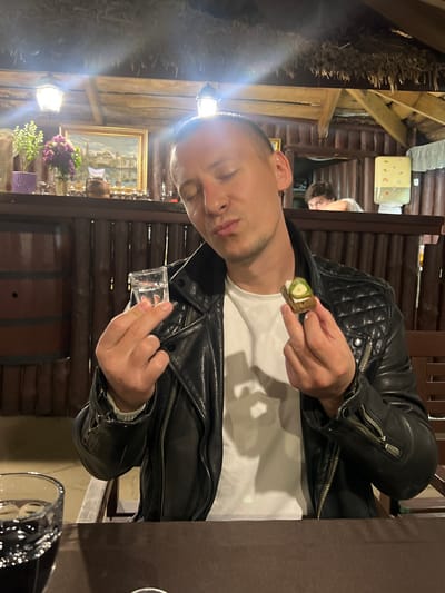 The image shows a man in a black leather jacket and white t-shirt, holding a piece of bread topped with what appears to be a slice of cucumber. In his other hand, he holds a small glass of liquid. He appears to be in a restaurant or dining area with wooden walls, a painting, flowers, and a wine barrel. Another person is visible in the background. A partial view of a wine glass on a table is also visible. The setting is potentially a restaurant in the city of Samarskoye, Russia.