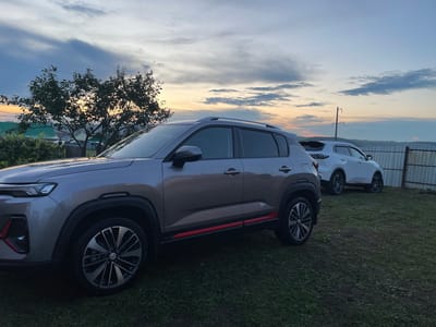 The photograph, taken at dusk in Yermekeyevo, Russia, captures an outdoor scene featuring two crossover SUVs parked on a grassy area. The foreground is dominated by a silver SUV, its left side facing the camera, showcasing its sleek design with dark rims and red accents on its lower body. Behind it, a white SUV is partially visible, also on the grass. The background is a gradient of twilight sky with scattered clouds, transitioning from warm sunset hues near the horizon to cooler blues overhead. A silhouetted tree with lush foliage stands to the left, partially obscuring a residential area with green roofs and some power lines. A metal fence runs along the right side of the frame. The overall atmosphere is serene, characteristic of a rural evening. No people are present in the image.