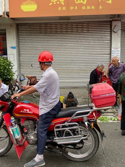 The image depicts a street scene in Chongzhou City, China, likely a market or commercial area, with a focus on a man on a red motorcycle. The man is wearing a red hard hat and a checkered shirt, and appears to be preparing to ride. In the background, several elderly individuals are visible, some standing and conversing, while another is kneeling on the ground. A shop with a rolled-down metal shutter is also prominent. The street appears to be paved, and there are some vegetables, possibly beans and watermelons, laid out on the ground near the people in the background. The overall atmosphere suggests a bustling, everyday scene in a Chinese city.