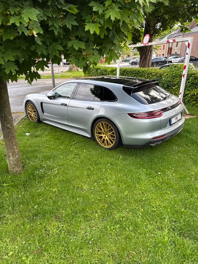 The image shows a silver Porsche Panamera Sport Turismo parked partially on a grassy area next to a sidewalk and tree in Melreux, Belgium. A height restriction bar, red and white, is overhead. Other cars are parked on the street in the background. A height limit sign of 2.2 meters is visible. The car has gold rims. The license plate is partially visible. The location is recognizable by the architectural style of the nearby buildings.
