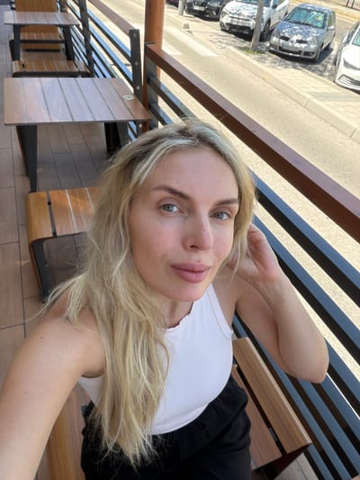 A blonde woman with light skin and fair complexion is seated on a wooden bench at an outdoor cafe. She is wearing a white tank top and black pants. Her hair is long and blonde, framing her face. She is looking at the camera and touching her hair. The background shows outdoor seating with wooden tables and chairs, and beyond that, a street with parked cars. The lighting suggests it is daytime.