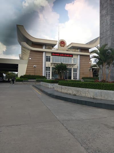 The image shows a view of the Moc Bai International Border Gate in Ben Cau, Vietnam, on a cloudy day. The border gate building is a modern structure with a beige facade and curved architectural elements. Above the entrance, signage reads "CỘNG HÒA XÃ HỘI CHỦ NGHĨA VIỆT NAM" (Socialist Republic of Vietnam) and "CỬA KHẨU QUỐC TẾ MỘC BÀI" (Moc Bai International Border Gate). A large, ornate pillar with bas-relief carvings stands to the right of the building. In front of the building, there are manicured green hedges and palm trees. The foreground consists of a concrete paved area with yellow lines marking lanes. A person wearing a dark outfit and a face mask is walking away from the camera on the left side of the paved area. To the left of the main building, a covered structure with a bus is visible. The sky is overcast with dramatic clouds.
